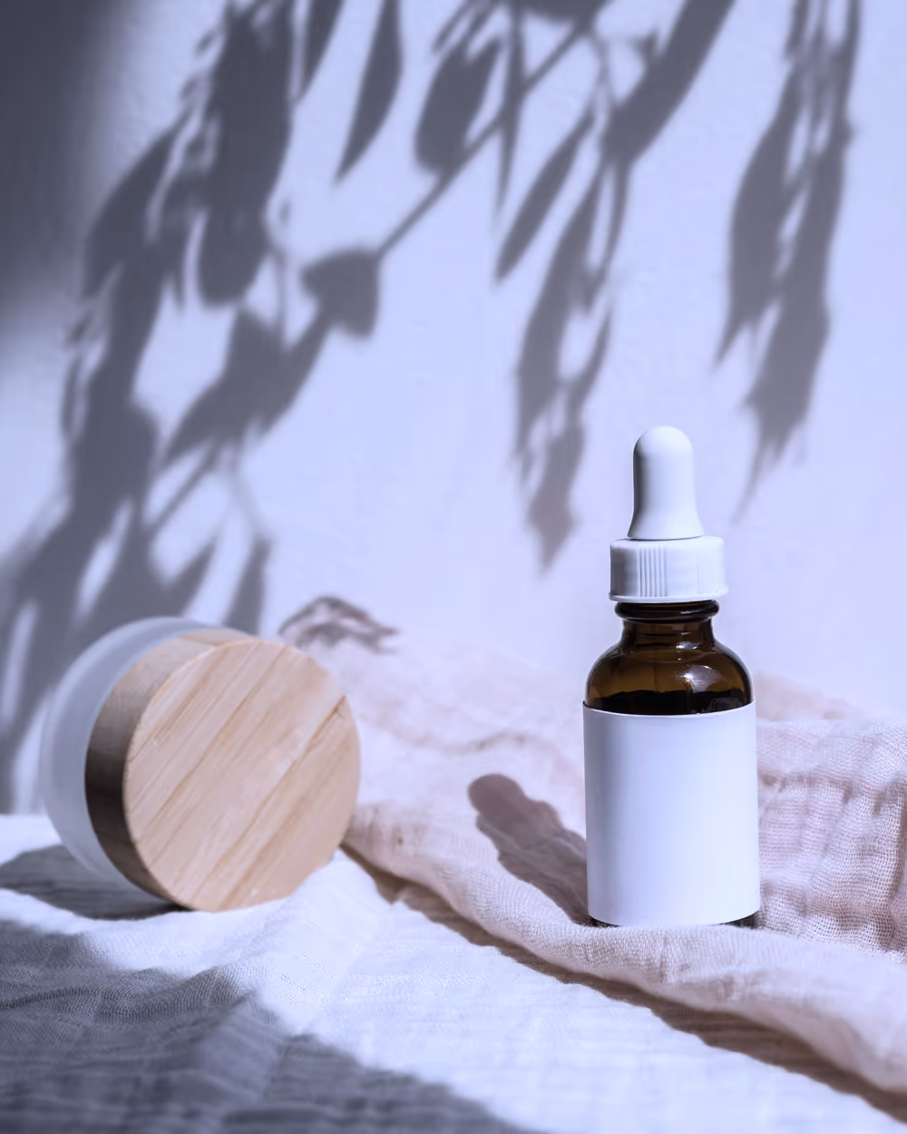 A dropper bottle of treatment serum resting on folded linen, soft daylight casting leaf shadows on the wall behind it
